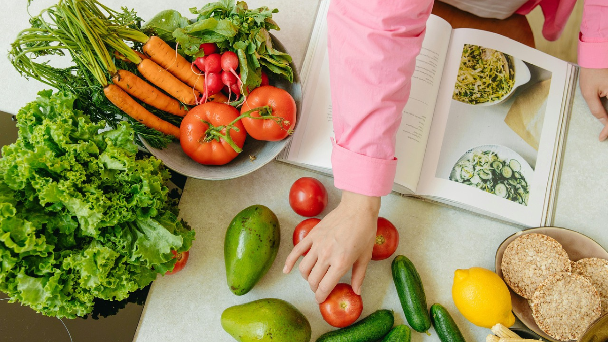 Pessoa folheando livro de receitas enquanto seleciona legumes frescos sobre a bancada, como cenouras, tomates, pepino, alface, abacates e limão.