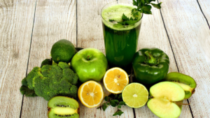 Um copo de suco verde rodeado por frutas e vegetais frescos, incluindo maçãs verdes, limões, abacate, brócolis, pimentão verde e kiwi.