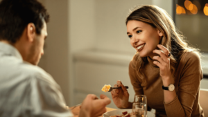 Mulher sorridente segurando um garfo com comida enquanto conversa com um homem durante um jantar romântico. Na mesa, há um prato de Strogonoff e uma taça, criando um ambiente acolhedor e intimista.