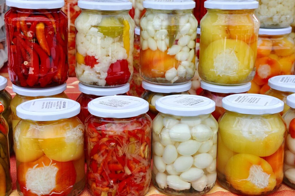 Diversos potes de vidro com conservas de vegetais coloridos, como pimentas, cebolinhas, couve-flor e pimentões.