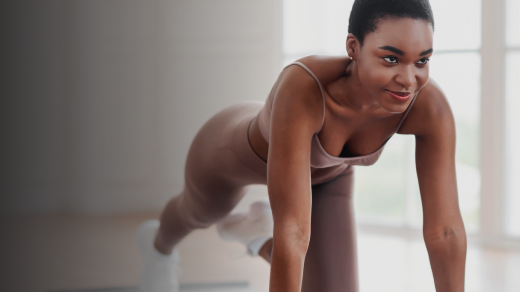Mulher com roupa de treino bege fazendo um exercício de escalador dentro de casa. Ela mantém uma expressão determinada e sorridente, transmitindo energia e motivação. O ambiente é bem iluminado, com fundo desfocado e janelas ao fundo.