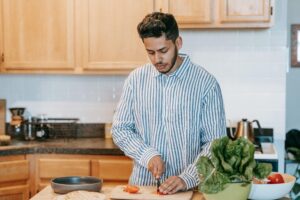 Esta imagem retrata um momento de preparo de refeição saudável em casa. O homem está cortando tomates frescos, enquanto ingredientes como alface e outros vegetais estão dispostos próximos, prontos para serem usados. Essa cena reforça a importância de cozinhar com ingredientes naturais, promovendo bem-estar, equilíbrio nutricional e autonomia alimentar.