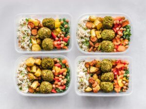 Esta imagem mostra quatro marmitas saudáveis e coloridas, compostas por arroz com ervas, legumes assados como batata e cenoura, grão-de-bico, pimentão e bolinhos de falafel. É um exemplo de refeição balanceada, rica em proteínas vegetais, fibras e nutrientes essenciais. Perfeita para quem busca praticidade sem abrir mão de uma alimentação nutritiva.