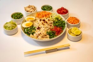 A imagem apresenta um prato de macarrão oriental cuidadosamente montado, com legumes frescos e coloridos, ovo cozido, frango desfiado e temperos típicos. Ao redor, pequenos recipientes contêm acompanhamentos e condimentos que permitem personalizar o sabor. É uma refeição equilibrada, nutritiva e inspirada na gastronomia asiática, perfeita para quem aprecia pratos leves e saborosos.