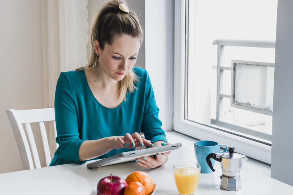 Mulher sentada à mesa com frutas e café da manhã, usando tablet e refletindo, representando momentos ligados à fome emocional