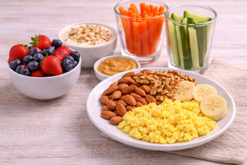 Prato com ovos mexidos, banana, amêndoas e nozes ao lado de frutas vermelhas, iogurte, pasta cremosa e palitos de legumes sobre mesa clara.
