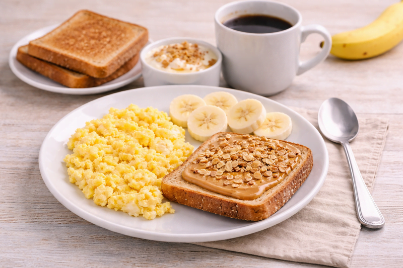 Café da manhã econômico com ovos mexidos, pão integral com pasta e aveia, rodelas de banana, iogurte e café sobre mesa clara.