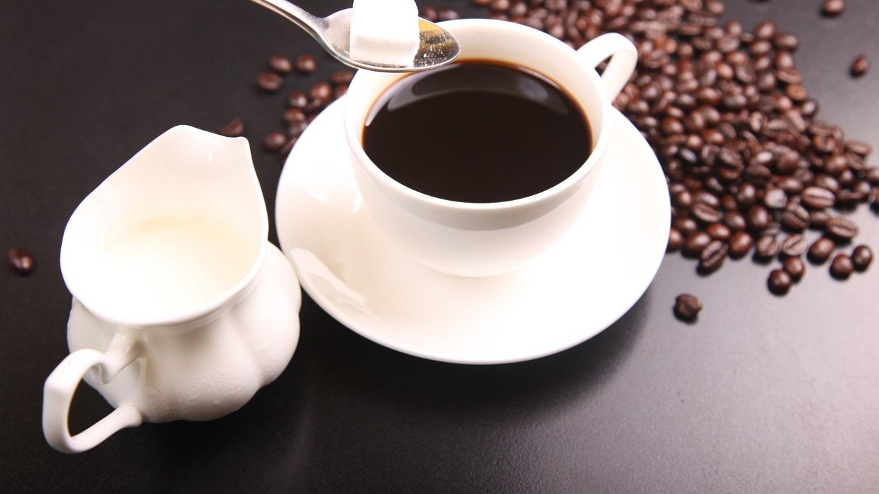Xícara de café preto com um cubo de açúcar sobre a colher, ao lado de uma jarra de leite