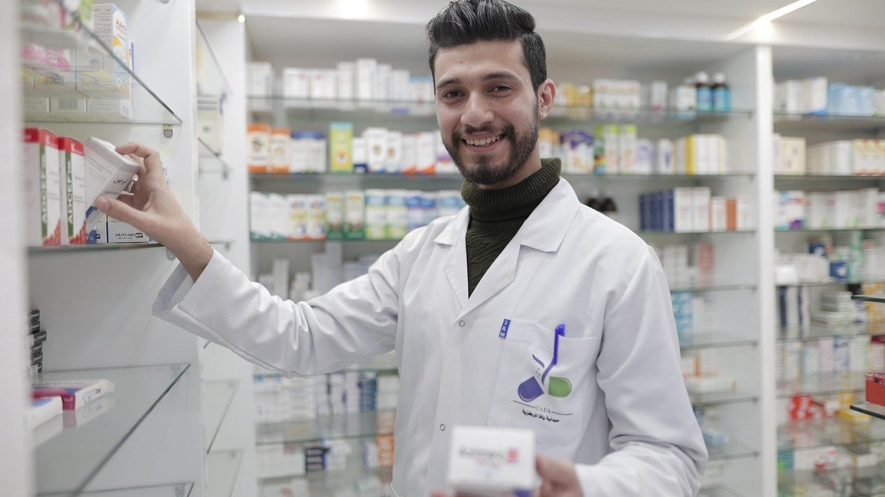 Farmacêutico sorrindo segura uma caixa de medicamento ao lado das prateleiras de uma farmácia