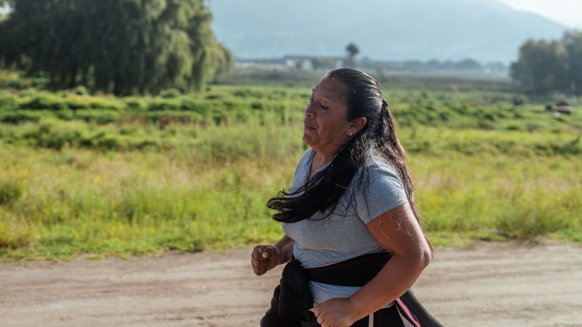 Mulher de cabelo preto longo correndo em uma estrada de terra, com campo e montanhas ao fundo