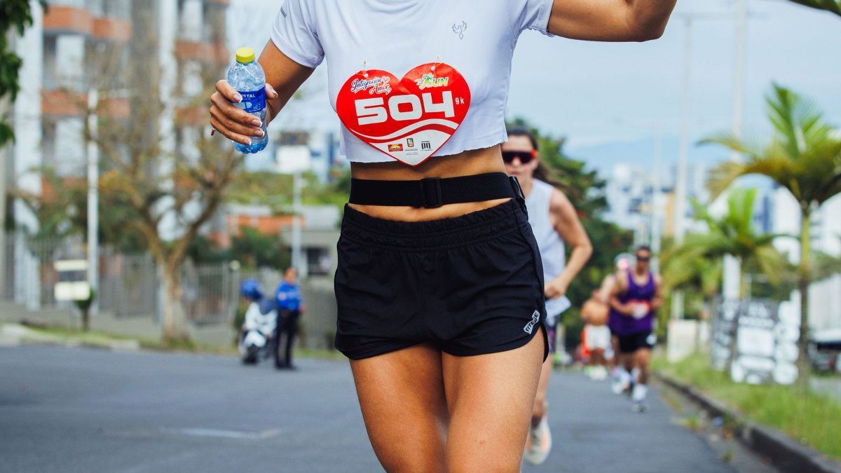 Corredora de camiseta branca com número de prova e short preto, com uma garrafa de água na mão, correndo em uma rua urbana