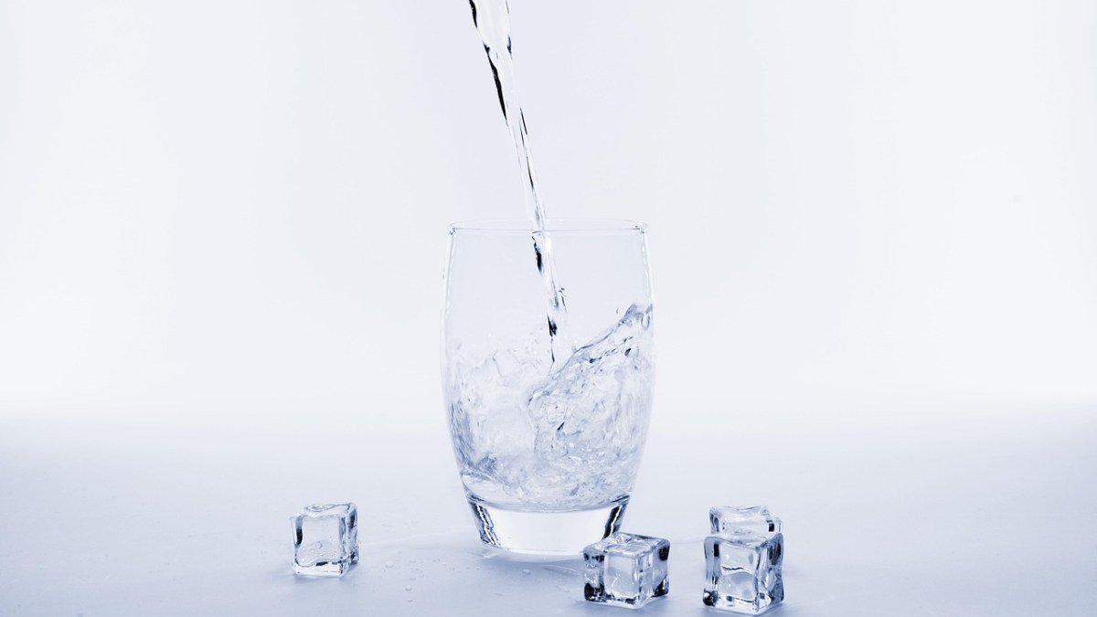 Água sendo servida em um copo transparente, com cubos de gelo ao redor