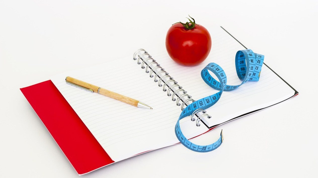 Caderno aberto com um tomate, fita métrica e caneta, para anotar a dieta e calcular as calorias do dia
