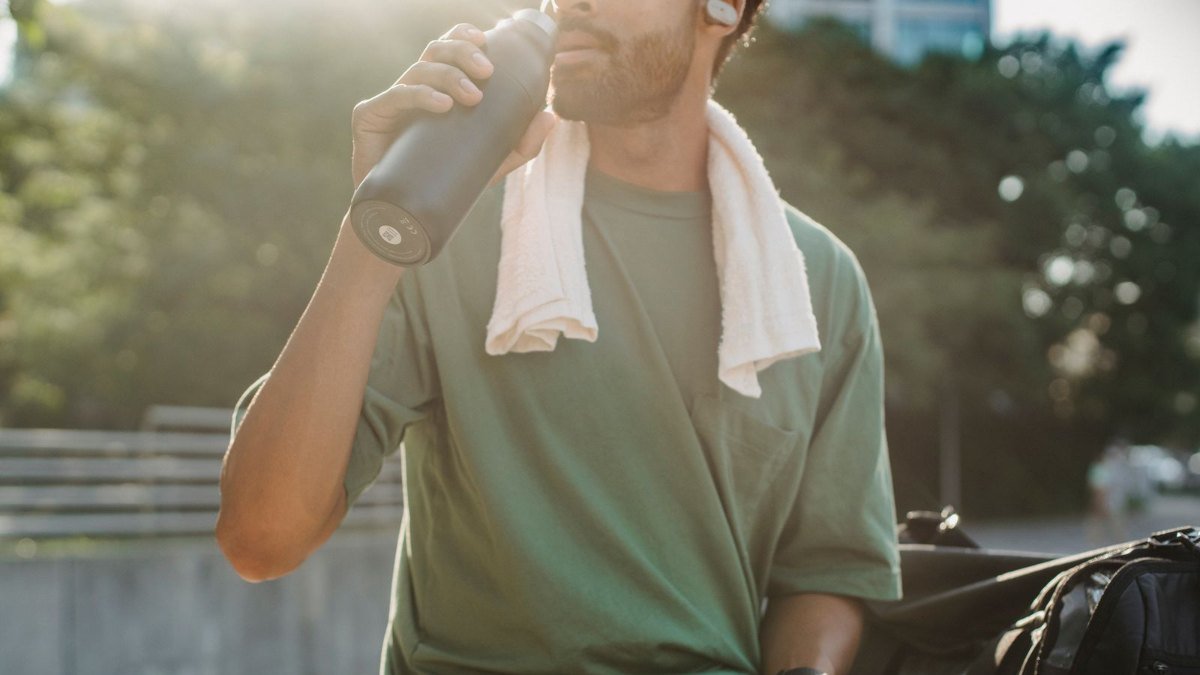 Homem de camiseta verde com toalha branca no pescoço bebendo água, reposição de líquidos para quem usa sauna para emagrecer