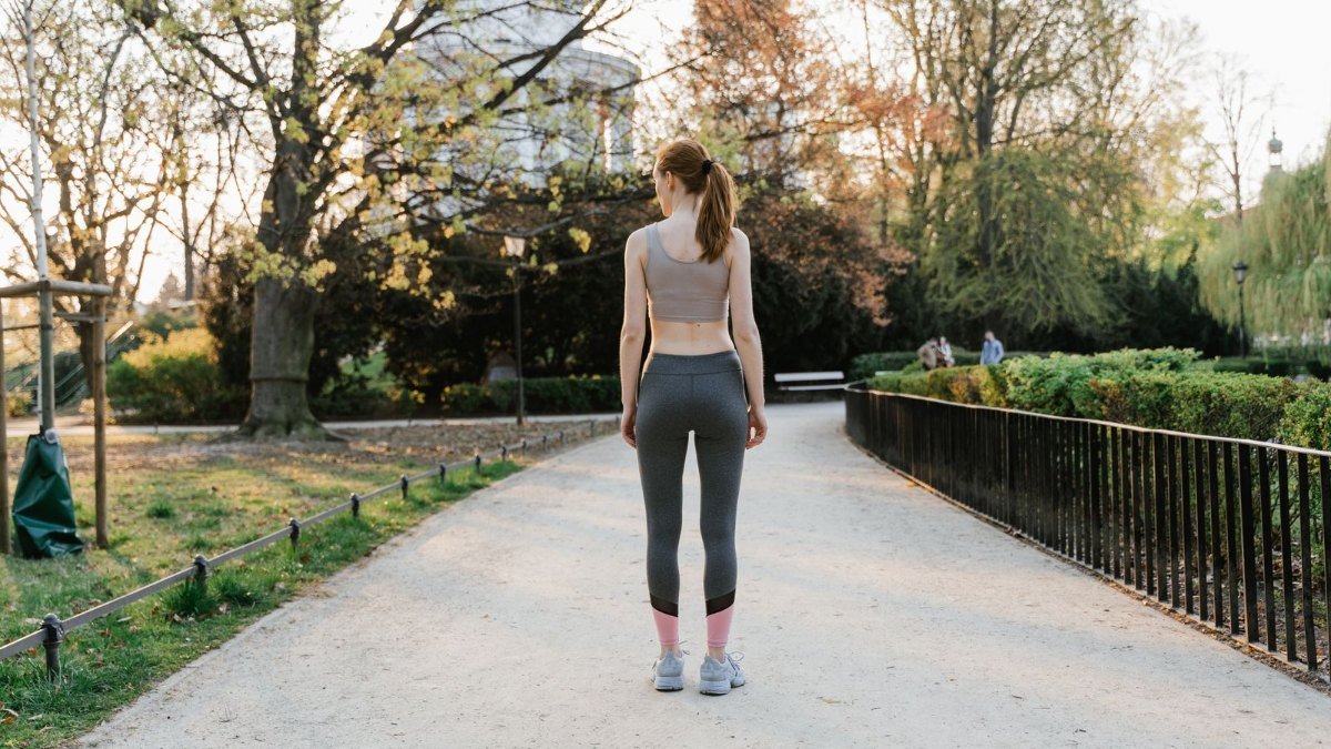 Mulher de roupa esportiva, de costas, em um caminho de parque cercado por árvores