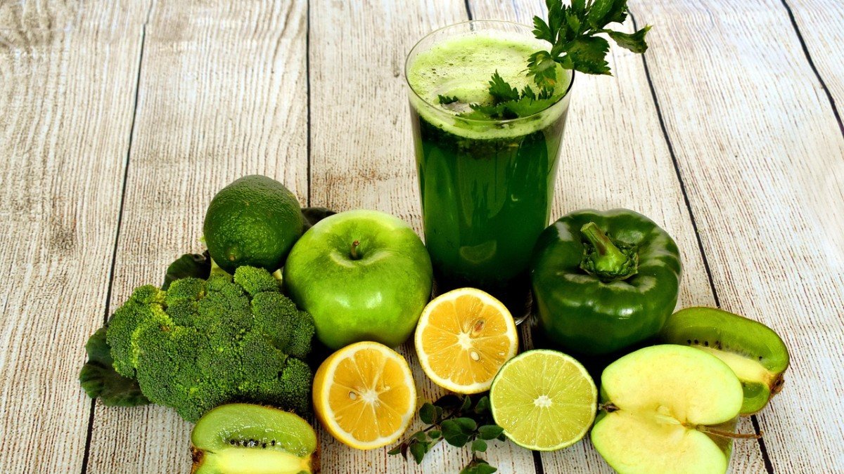 Copo de suco verde ao lado de brócolis, maçã verde, pimentão, limão, kiwi e maçã partida sobre mesa de madeira clara