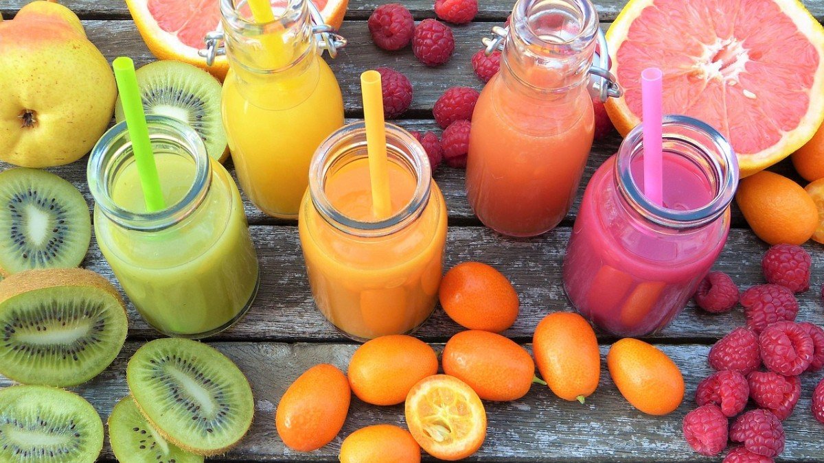 Sucos coloridos em potes de vidro com frutas Potes de vidro com sucos nas cores verde, amarelo, laranja e rosa, cercados de kiwi, laranja e framboesas
