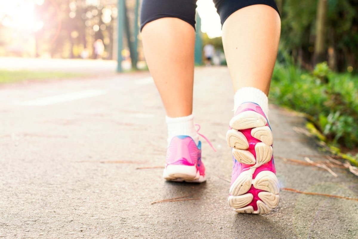 Pés de uma mulher caminhando em um caminho de parque, com tênis rosa e meias brancas, vistos por trás
