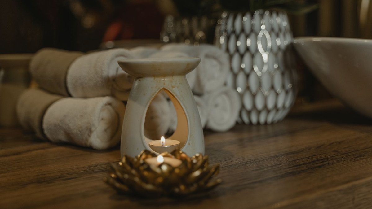 Toalhas brancas enroladas, uma vela acesa em um difusor de cerâmica e um vaso sobre uma bancada de madeira em um ambiente de spa